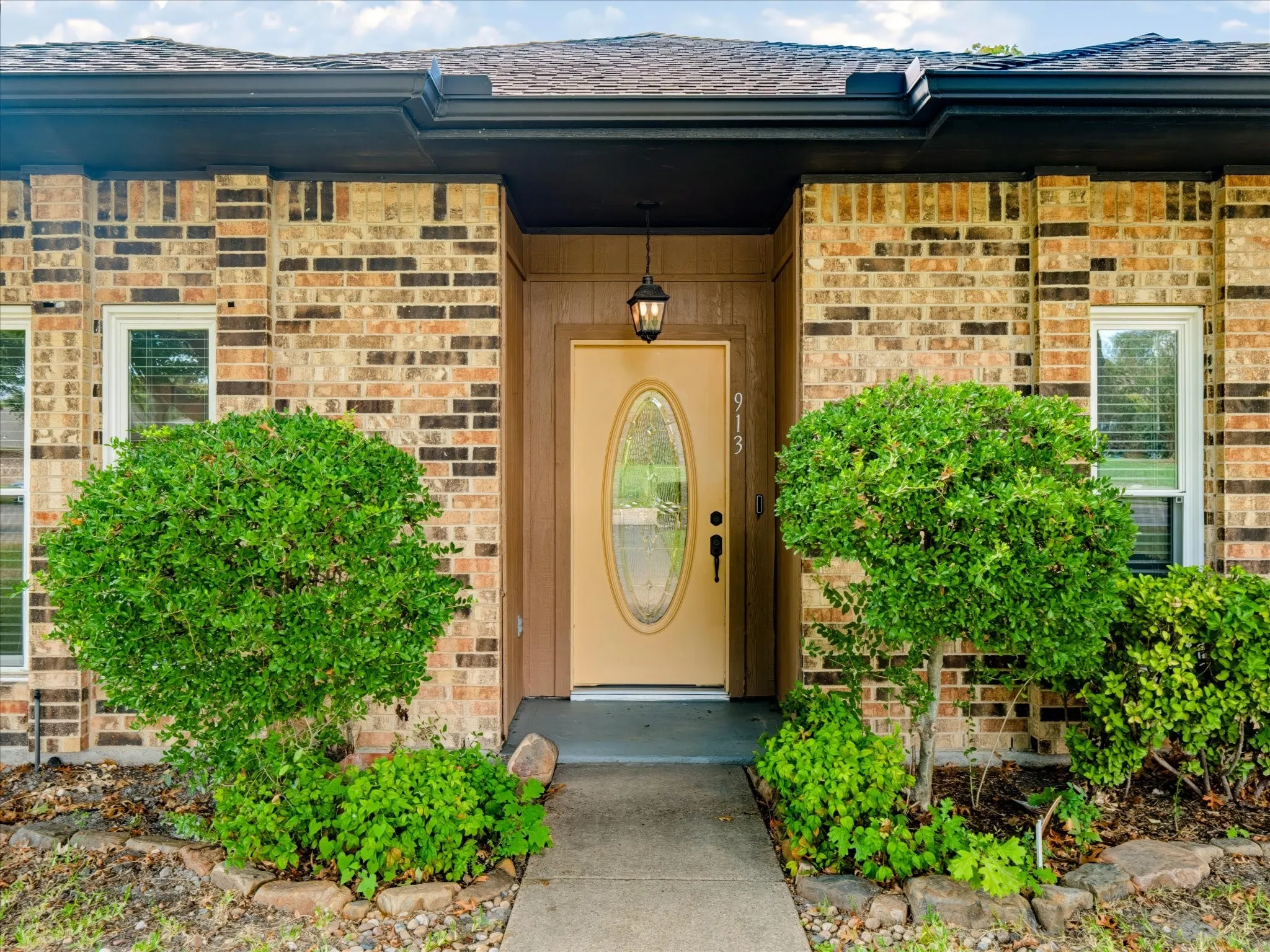 Brick exterior facade featuring a recessed entryway, a solid door with a central oval glass insert, a traditional hanging lantern fixture, and trimmed landscaping