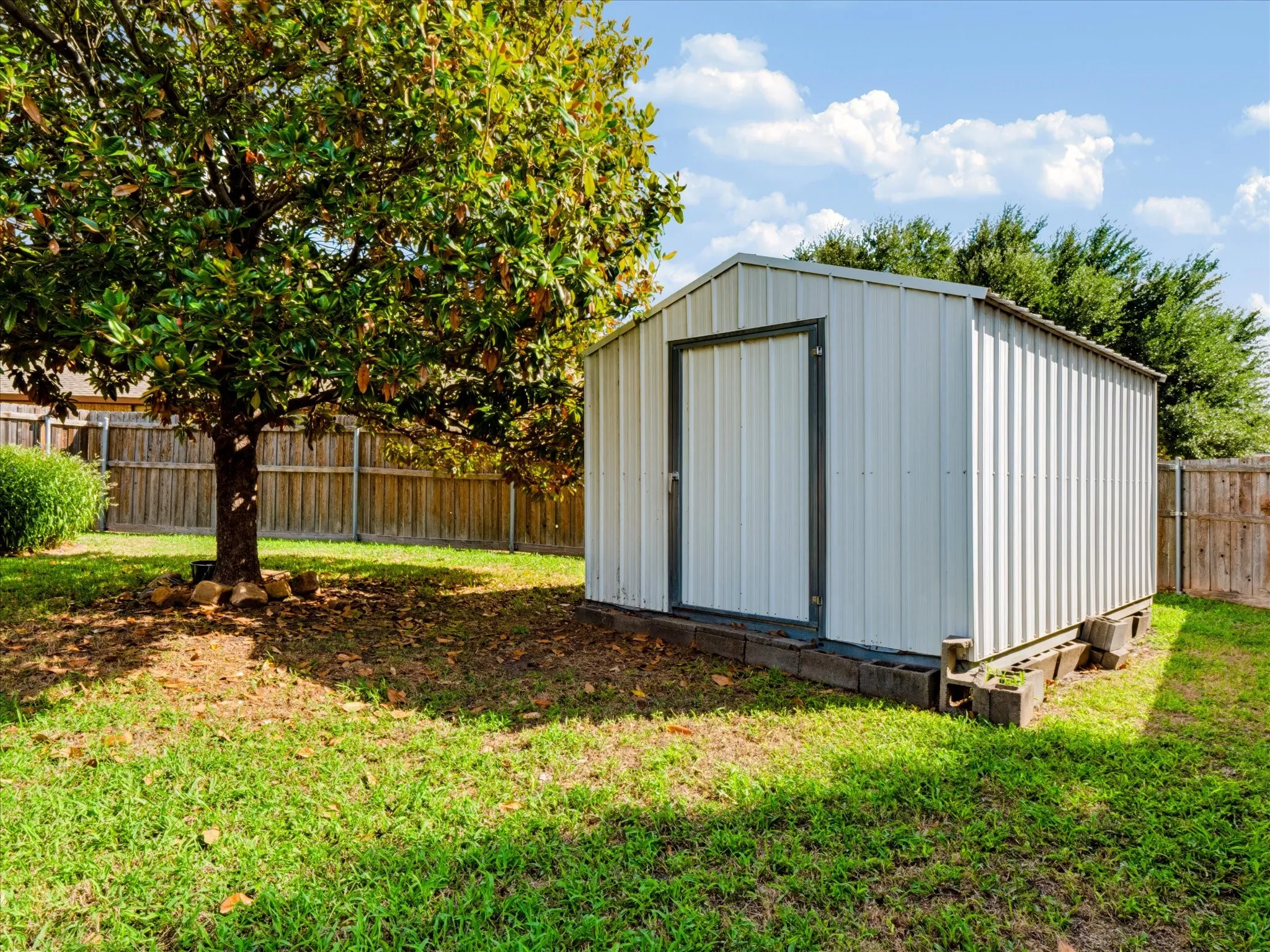 Metal storage shed with vertical siding, concrete masonry unit foundation, mature tree, and perimeter wood privacy fencing