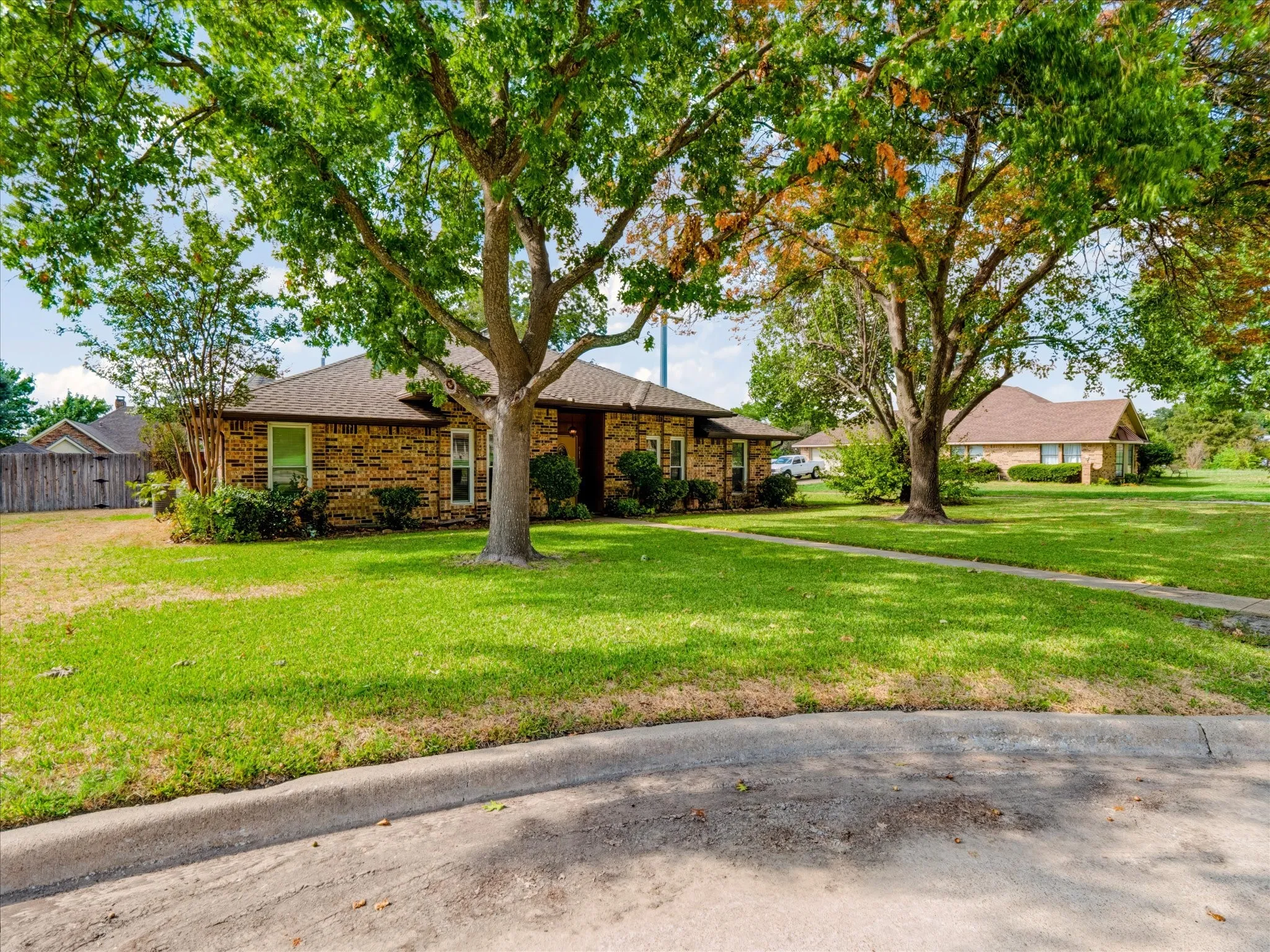 Brick exterior residence featuring a hipped roof, concrete walkway, and mature trees situated on a corner lot