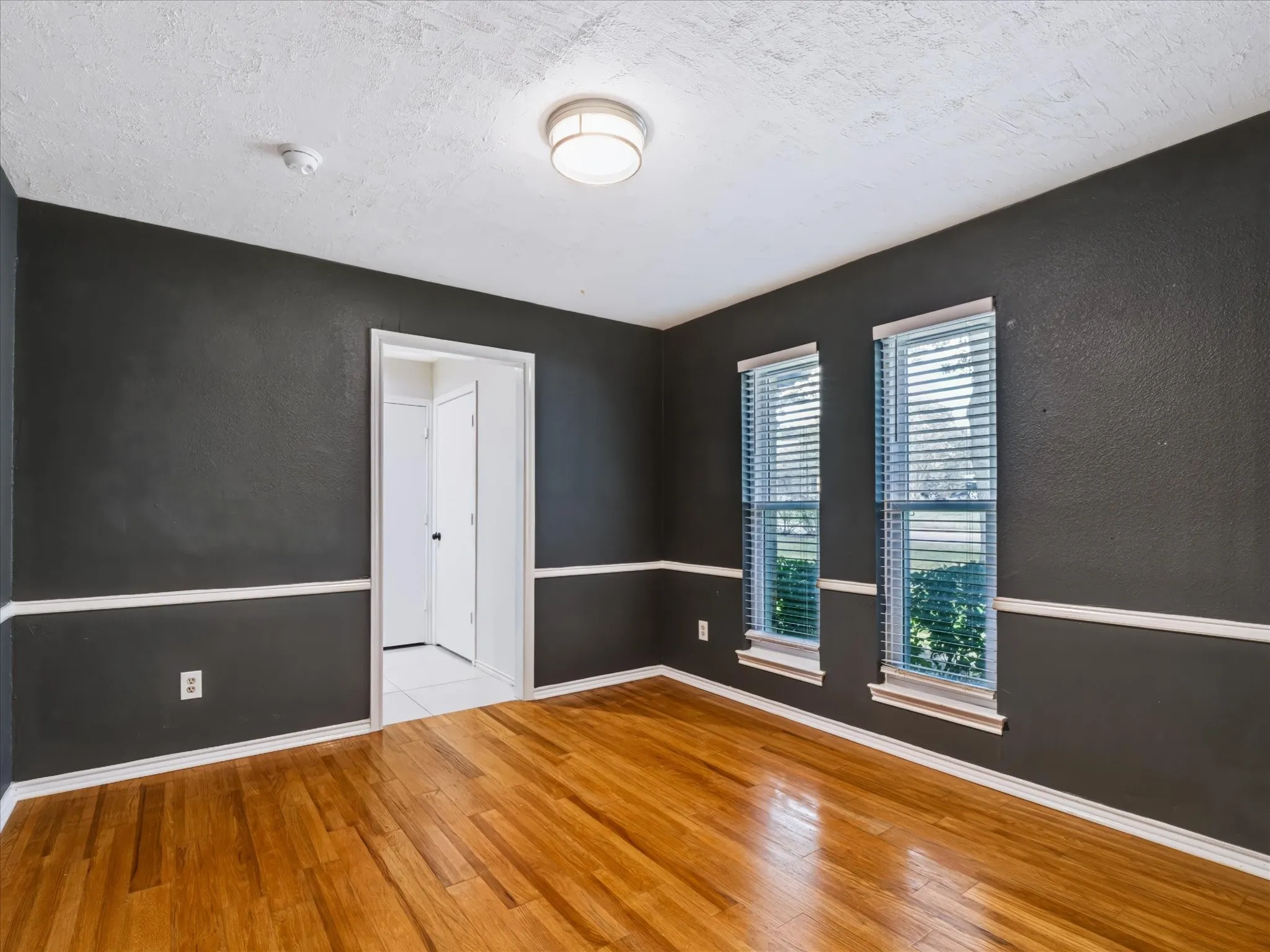 Room featuring wood-finish flooring, dark-toned walls with contrasting white chair rail molding, dual windows with integrated blinds, and a circular flush-mount ceiling fixture