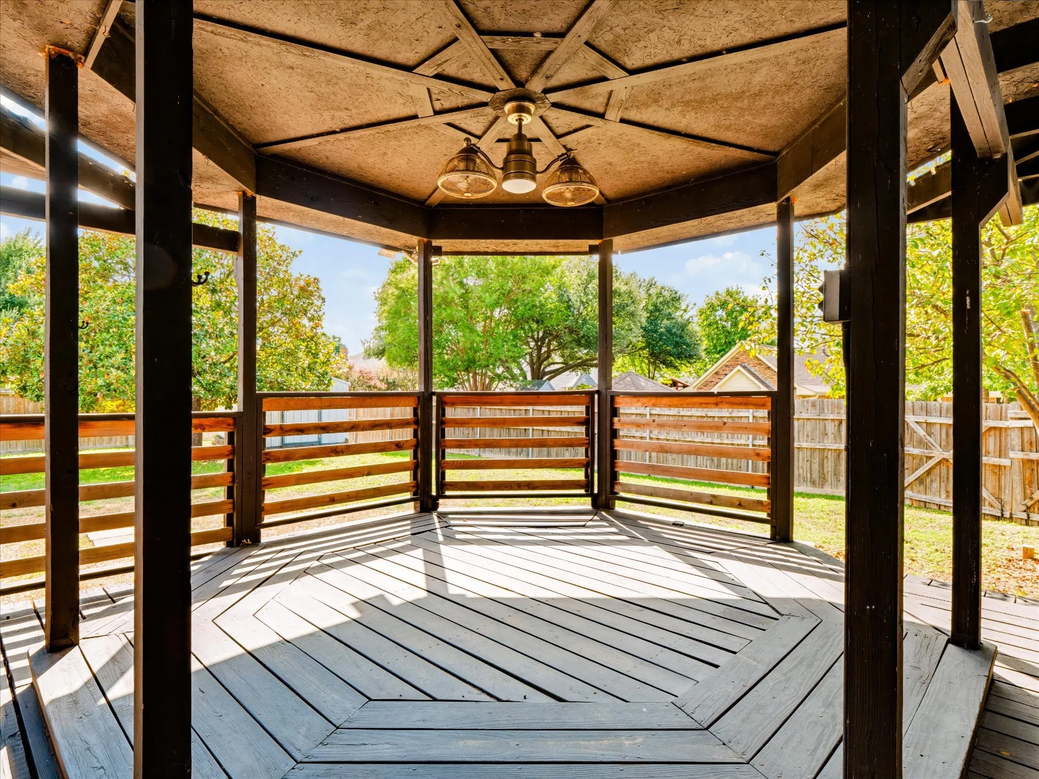 Octagonal wooden gazebo featuring a central multi-light pendant fixture, horizontal slat railing, and radial decking