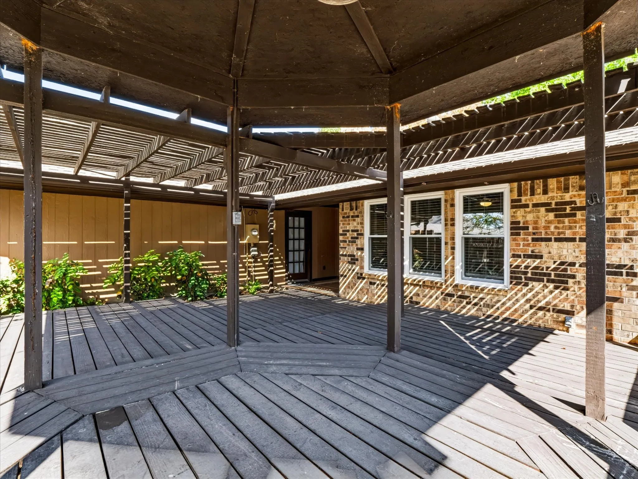 Covered wooden deck featuring a multi-faceted pergola, brick exterior siding, and three grid-pane windows