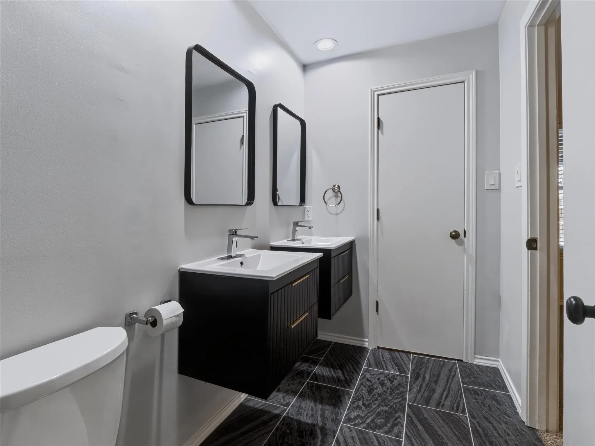 Dual floating vanities featuring matte black textured cabinetry, white integrated sinks, metallic hardware, and rectangular mirrors, complemented by large-format patterned stone-finish floor tiling