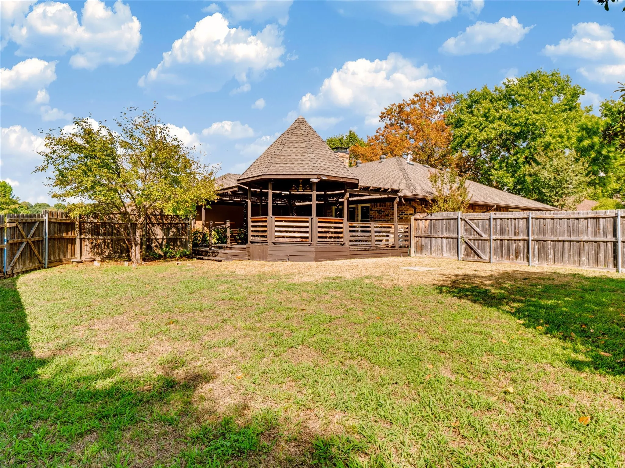 Expansive backyard featuring a leveled grass lawn, wooden perimeter fencing, mature trees, and a raised wooden deck with a gazebo-style roof structure