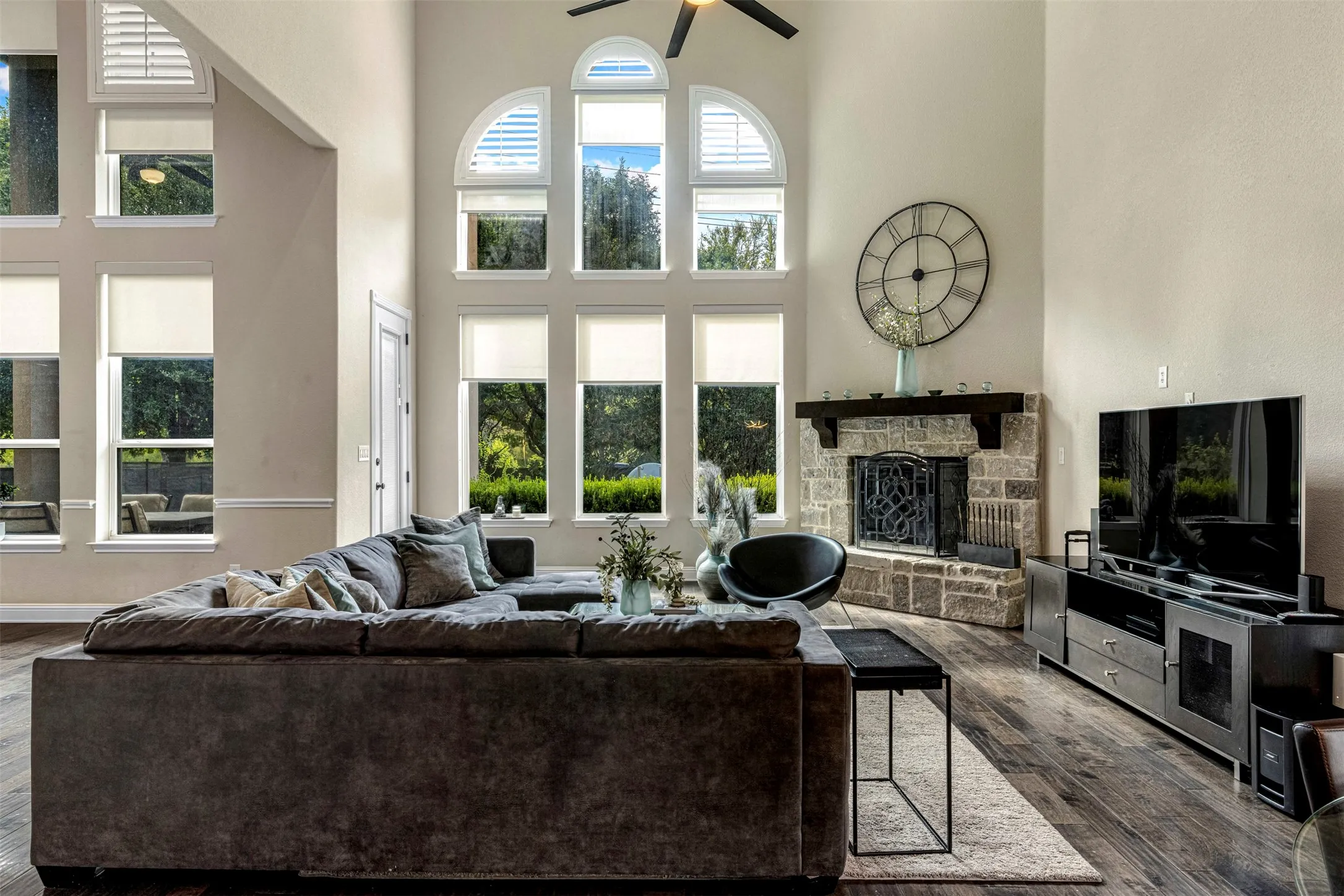 Living area featuring vaulted ceilings, floor-to-ceiling windows with arched transoms, stone-faced fireplace with a wood mantel, and wood-finish flooring