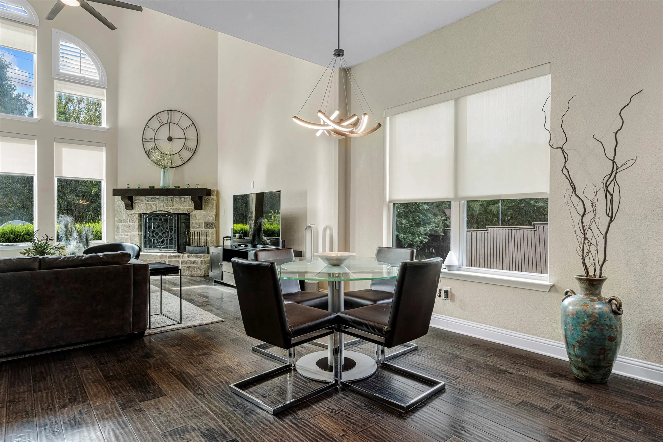 High-ceilinged living area featuring wood-finish flooring, a floor-to-ceiling stone-faced fireplace with a timber mantel, expansive arched windows, and a modern sculptural chandelier