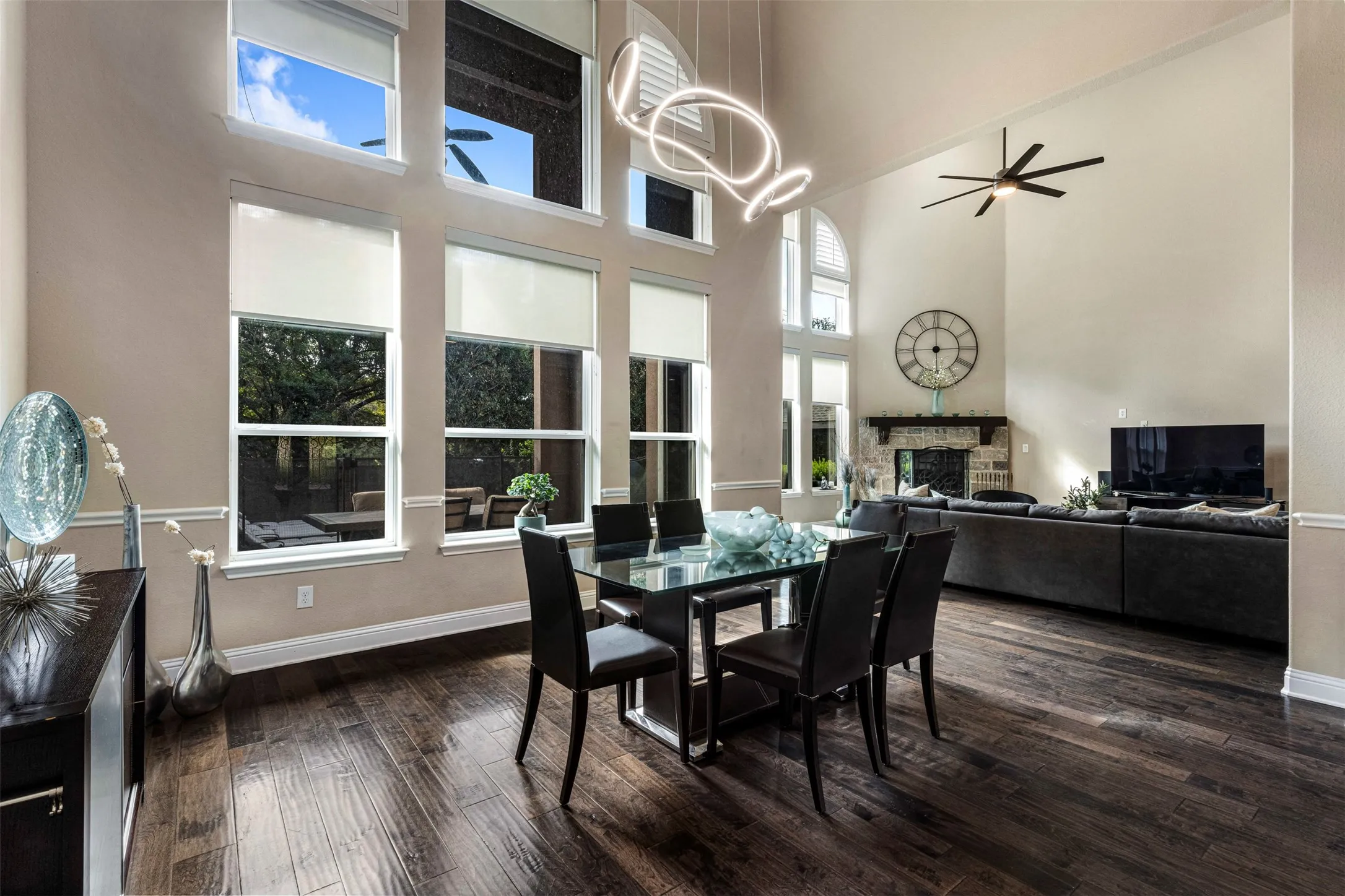 Vaulted ceilings featuring floor-to-ceiling multi-pane windows, wood-finish flooring, a stone-surround fireplace with a wooden mantel, and a contemporary architectural light fixture