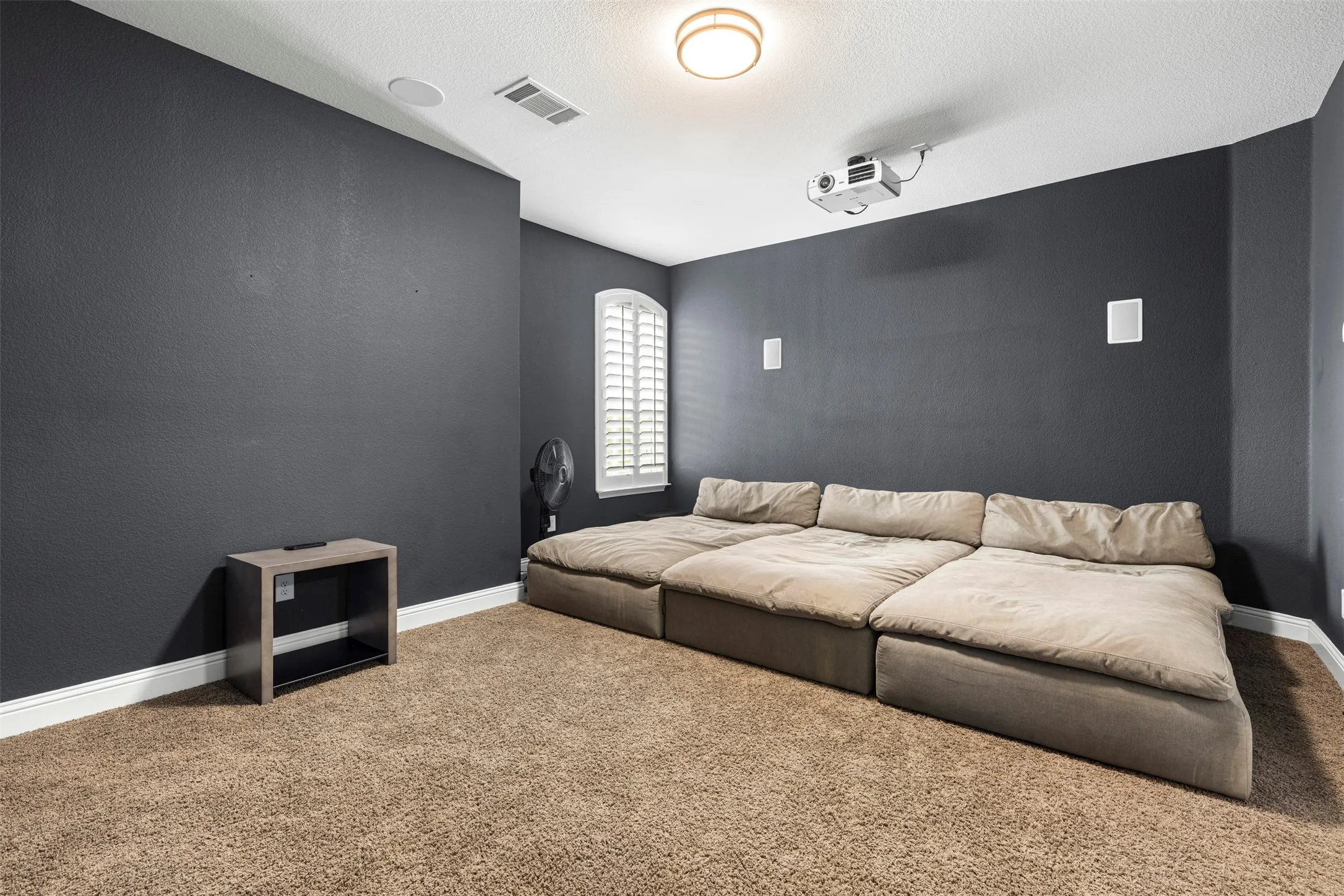 Room featuring wall-to-wall carpeting, ceiling-mounted projector, built-in wall speakers, and an arched window with plantation shutters