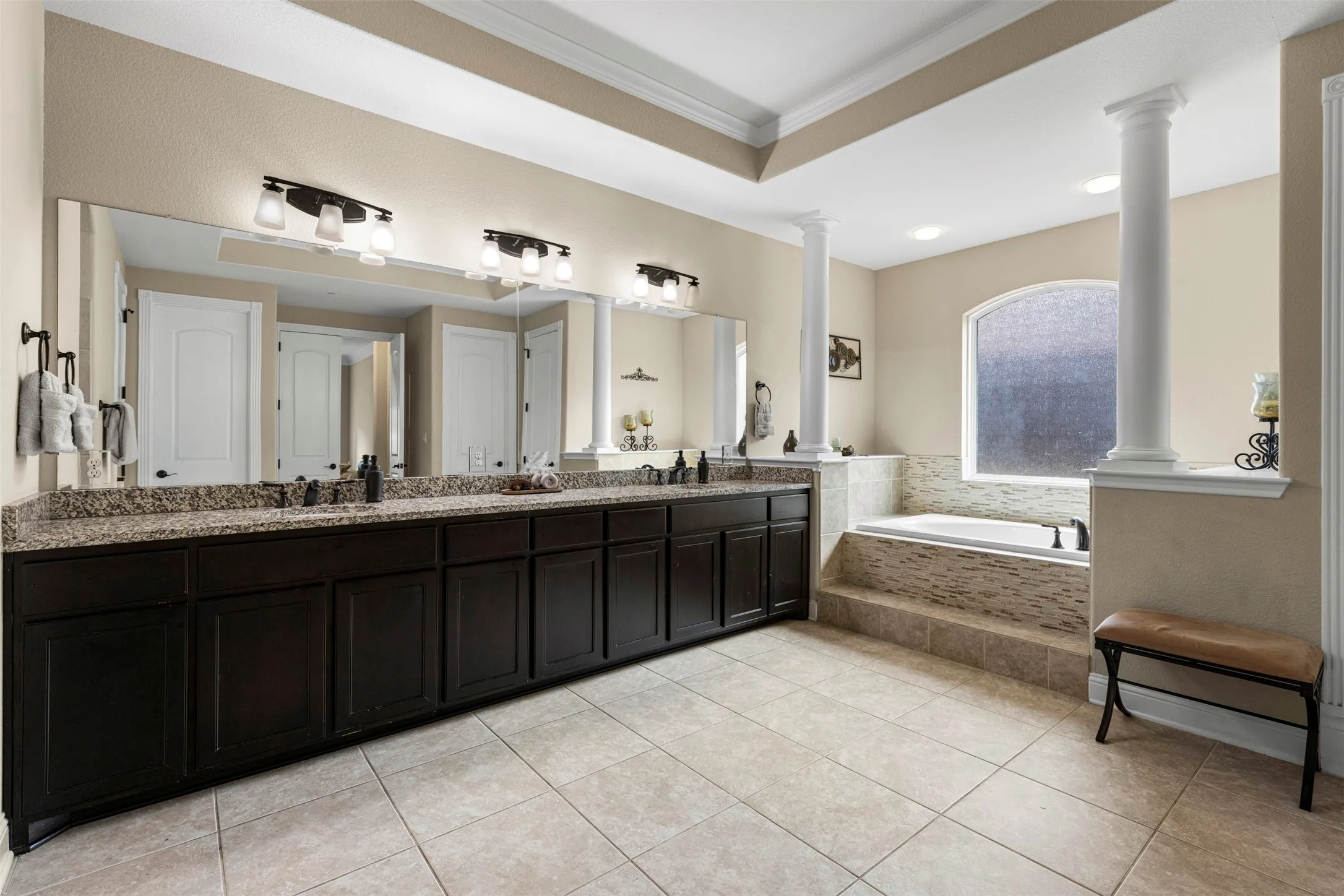 Double vanity with stone countertop, dark wood-finish cabinetry, built-in soaking tub with tile surround, arched window, and decorative architectural columns