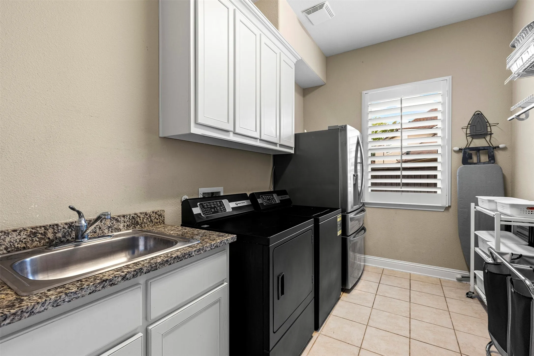 Utility room featuring white cabinetry, stone-finish countertops, stainless steel drop-in sink, tile flooring, and louvered window shutters