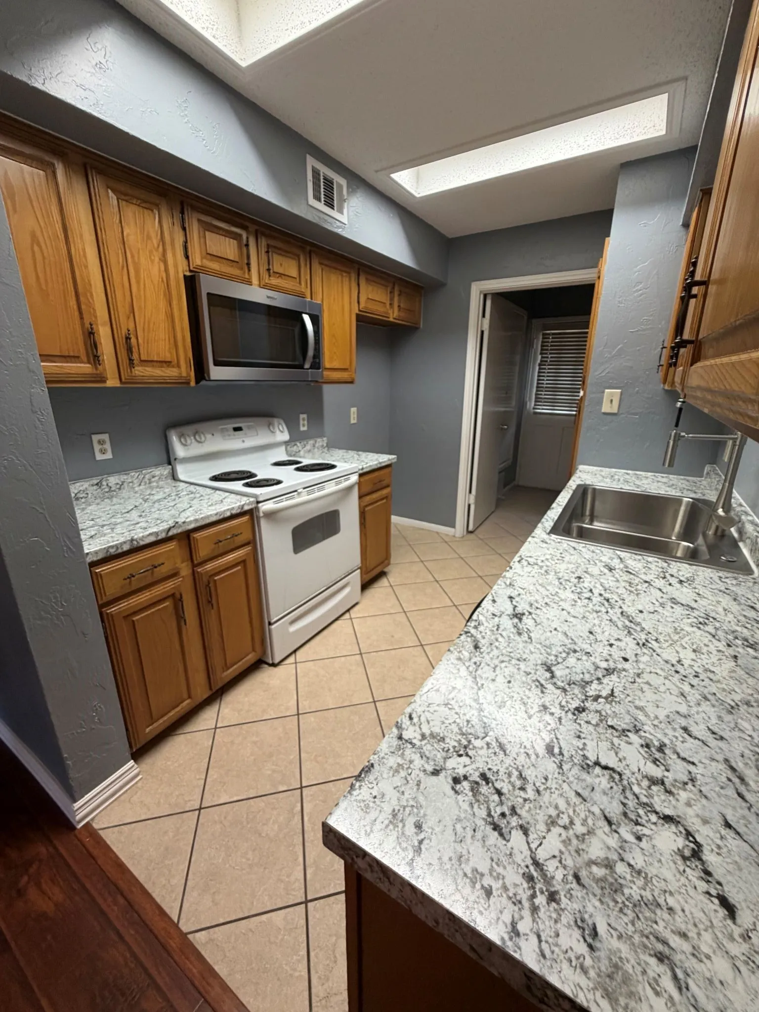 Kitchen featuring stone-finish countertops, wood-finish cabinetry, stainless steel sink with high-arc faucet, integrated microwave, and overhead skylight