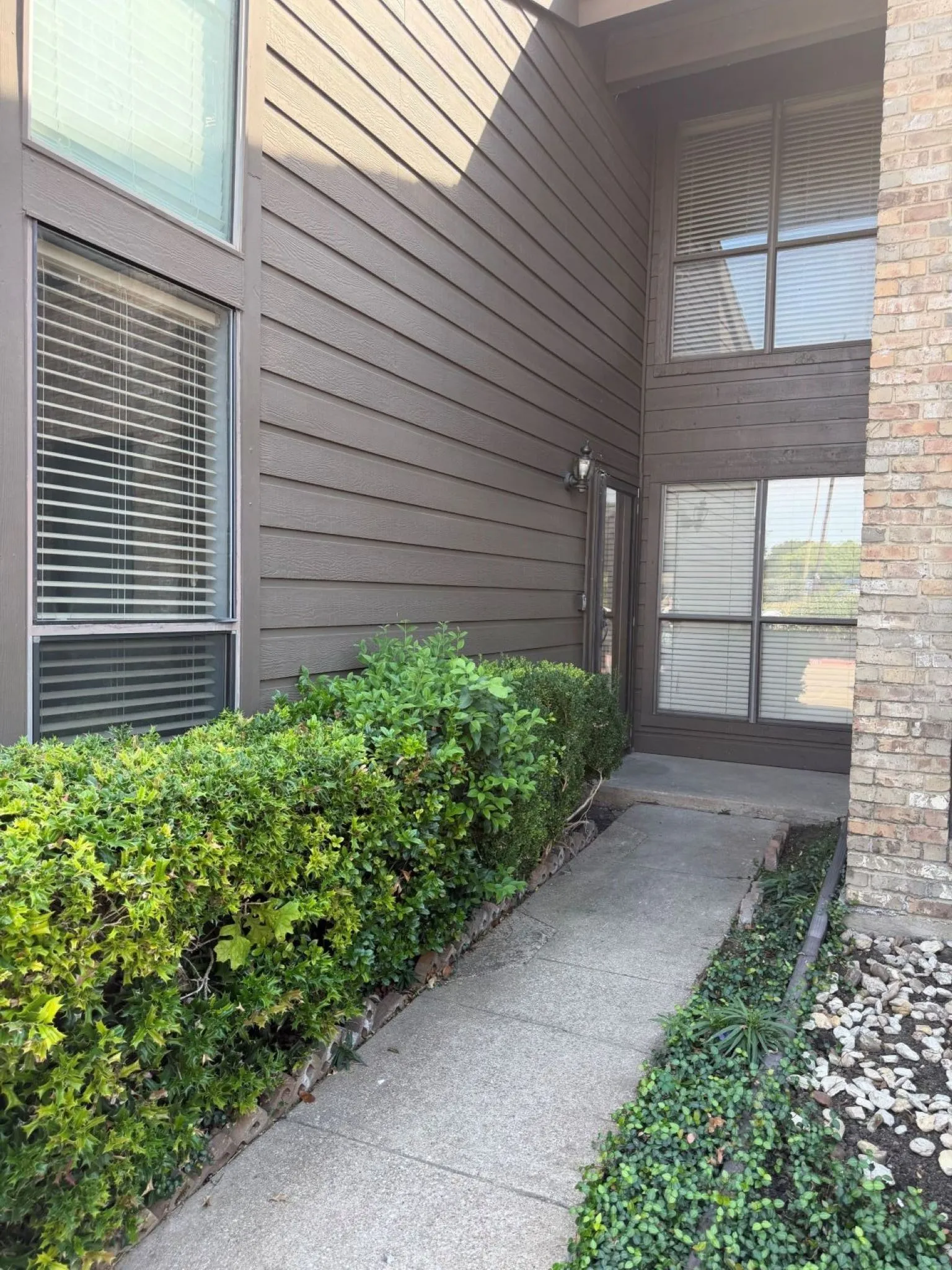 Exterior facade featuring horizontal siding and brick masonry, multi-pane windows, and a concrete walkway bordered by manicured shrubs