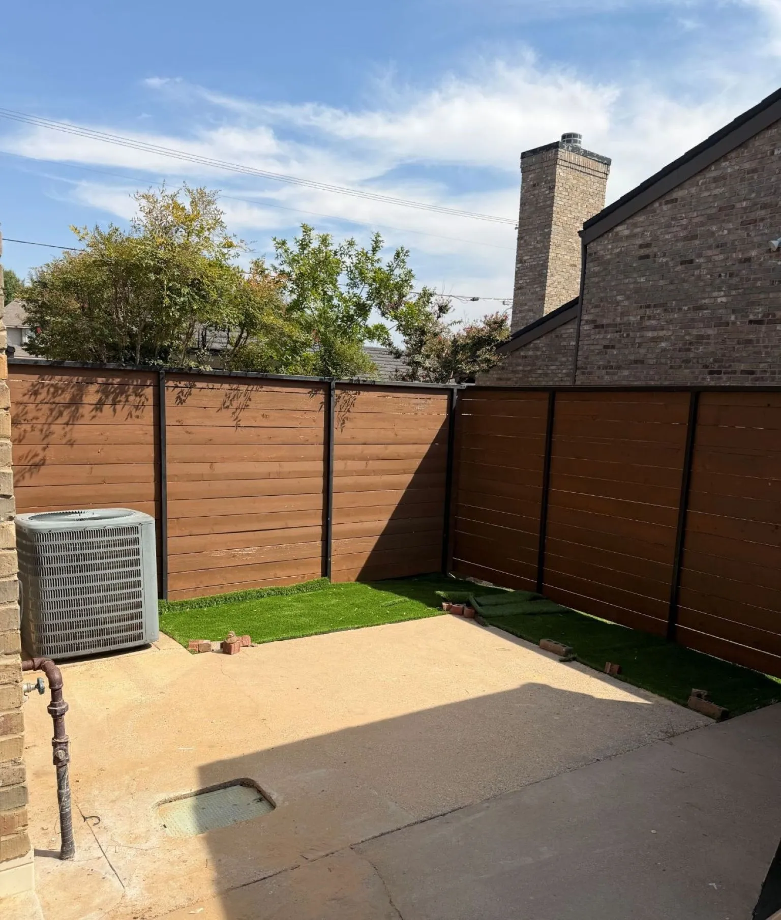Outdoor space featuring horizontal wood-slat privacy fencing, concrete patio, synthetic turf, and brick exterior chimney