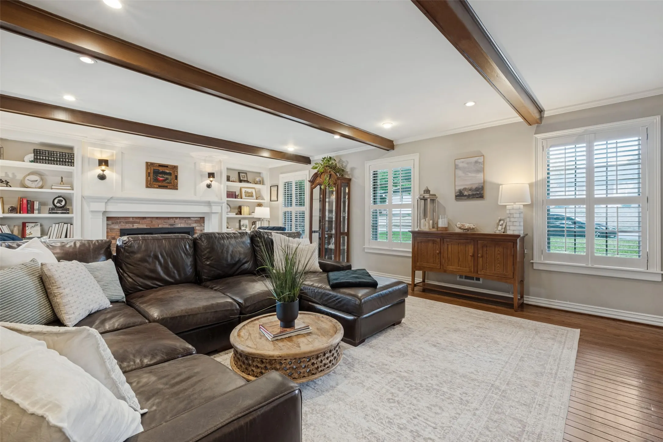 Living area featuring dark stained exposed ceiling beams, wood-finish flooring, built-in floor-to-ceiling bookshelves, and a brick fireplace surround