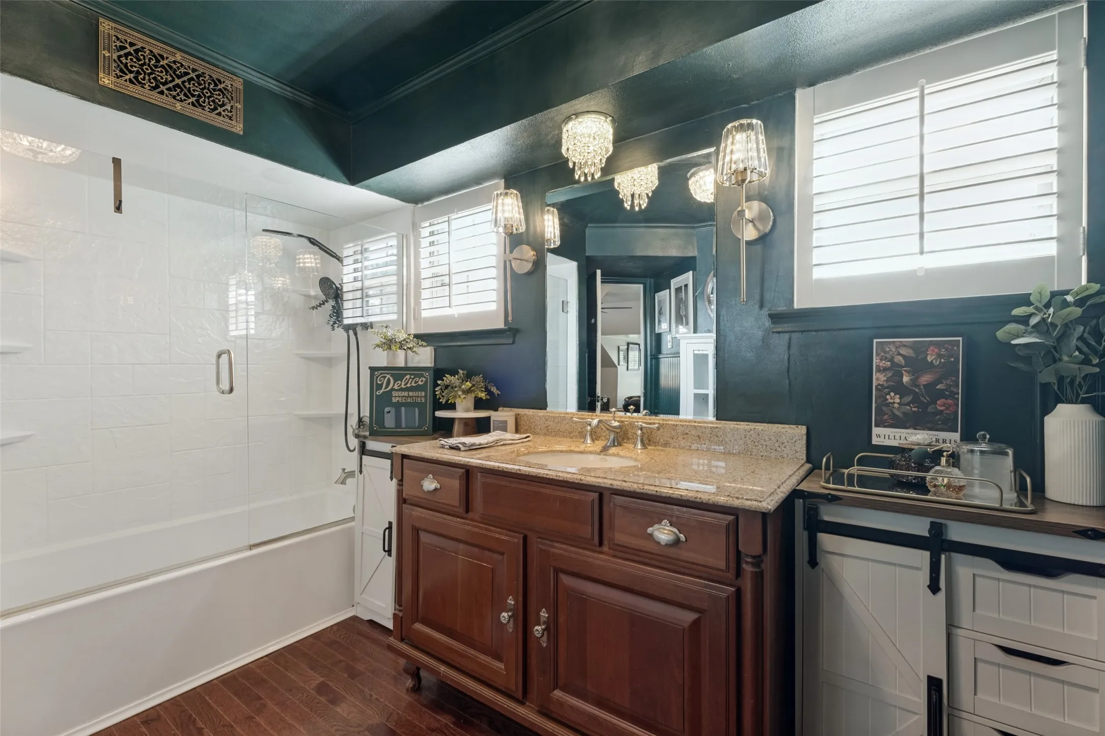 Wood-finish vanity with a stone countertop, white sliding-door cabinetry, wood-finish flooring, glass-enclosed shower, and plantation shutters