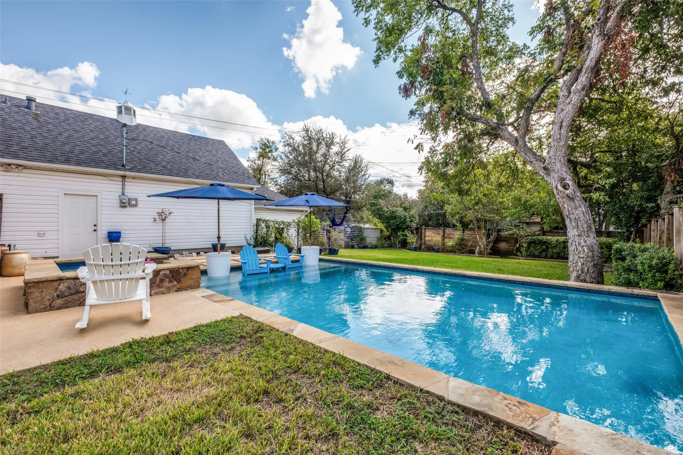 In-ground swimming pool featuring stone coping, integrated raised stone spa, concrete hardscaping, and mature trees