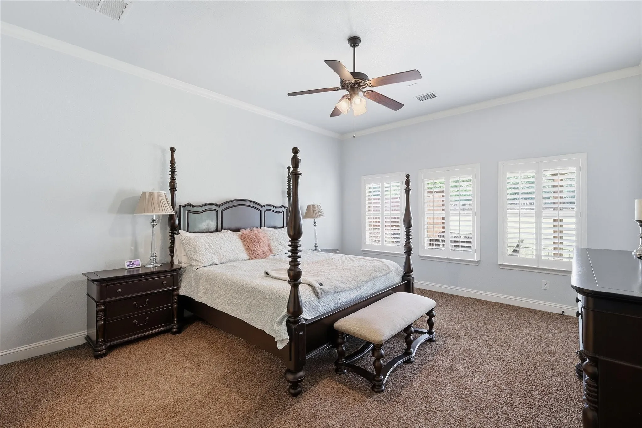 Spacious primary bedroom featuring neutral wall color, crown molding, wall-to-wall carpeting, triple shutter-covered windows, and a ceiling fan with integrated lighting