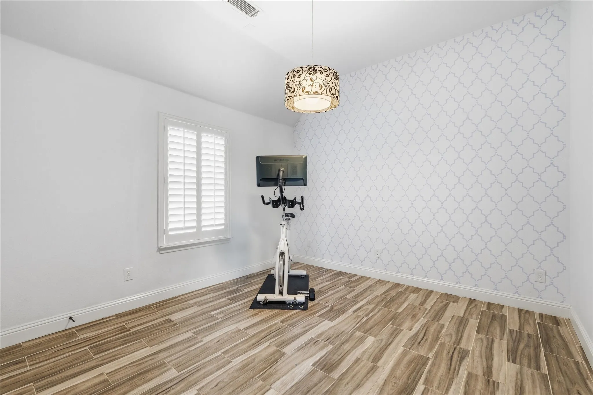 Wood-finish flooring, decorative patterned wallpaper, and a central drum pendant light fixture with laser-cut detailing, featuring a window with interior plantation shutters