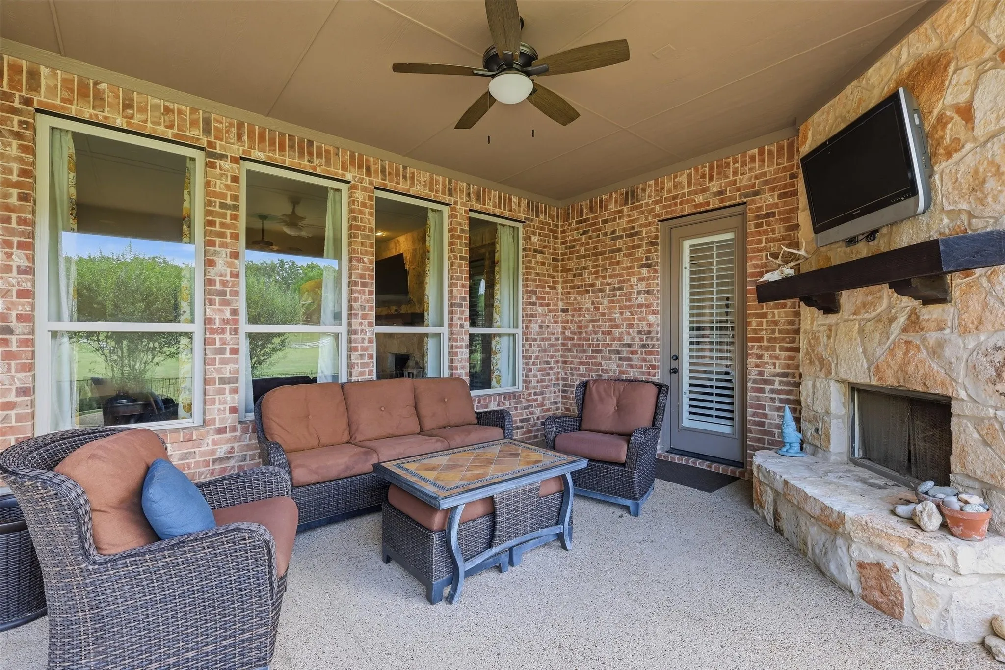 Covered patio featuring red brick walls, stone-surround masonry fireplace, timber mantel, and ceiling fan