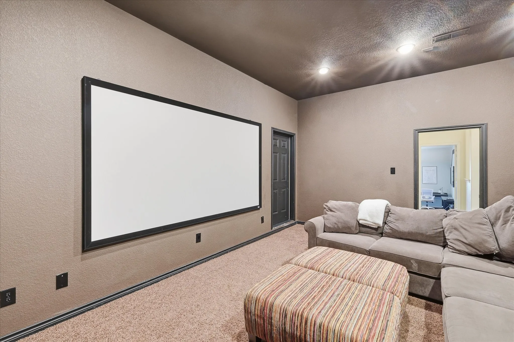 Dedicated media room featuring a mounted projection screen, recessed ceiling lighting, textured wall finish, and neutral carpeting