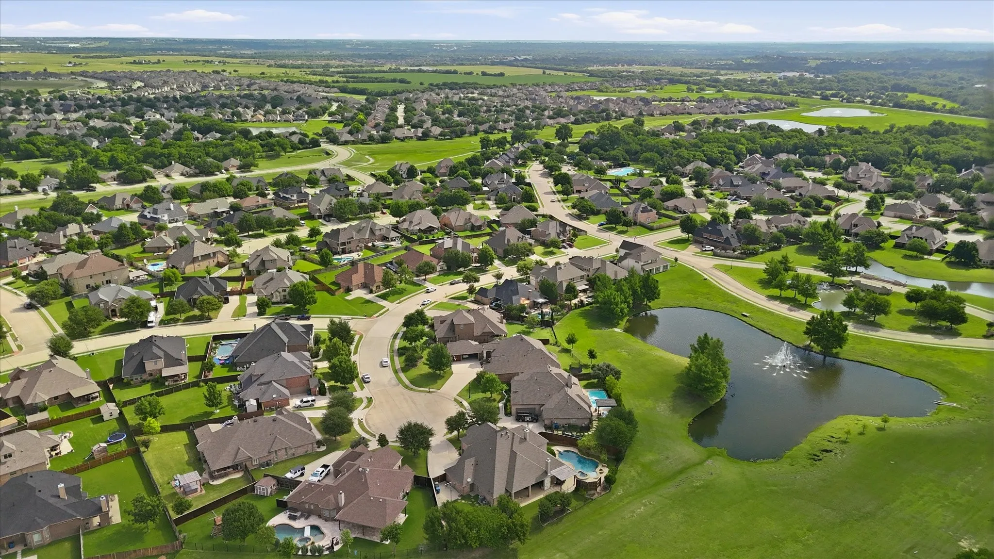 Aerial perspective of a residential community featuring expansive greenbelts, a decorative pond with a water feature, and mature trees