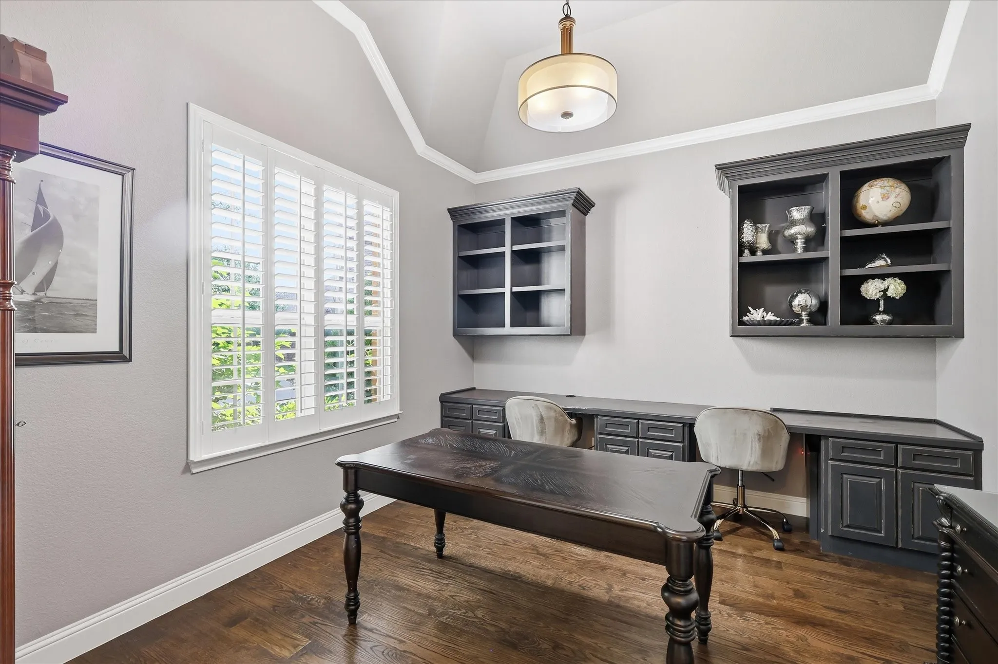 Vaulted ceiling featuring a central pendant light fixture, dark wood-finish flooring, plantation shutters, and extensive built-in dark cabinetry with integrated desk workspace