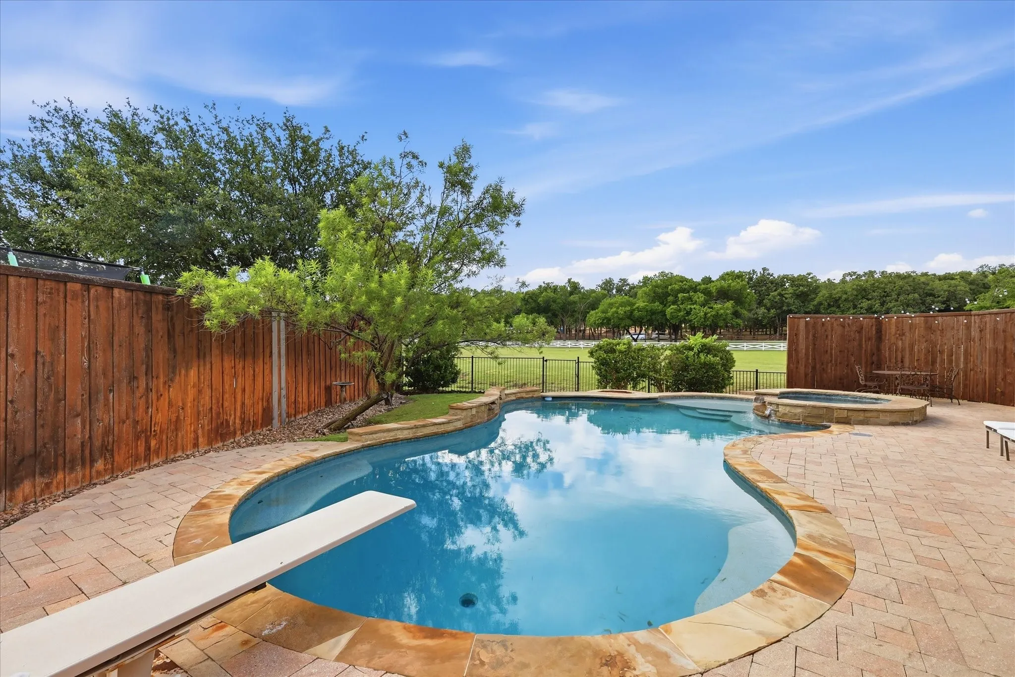 Backyard featuring an inground swimming pool with a diving board, elevated spa, natural stone coping, and expansive paver patio enclosed by a wood privacy fence
