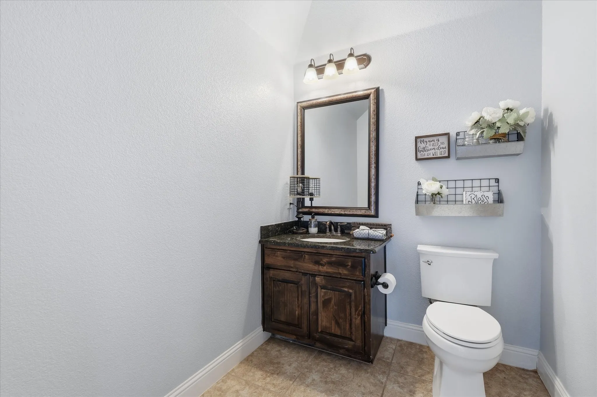 Full guest bathroom featuring a dark wood-finish vanity, stone-finish countertop with an undermount sink, framed wall mirror, three-light vanity fixture, and tile flooring
