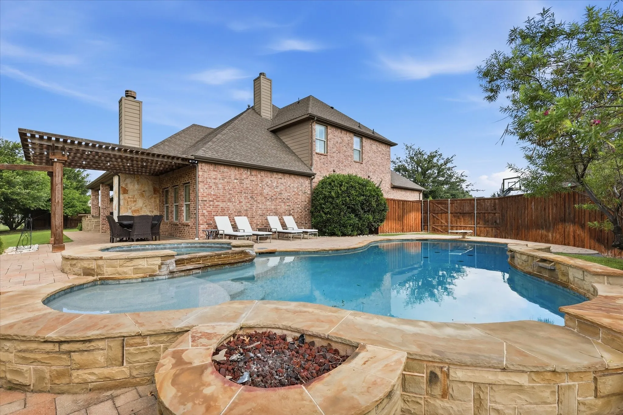Brick exterior home featuring a stone-clad swimming pool with integrated spa, built-in stone fire pit, covered pergola, and patterned paver patio