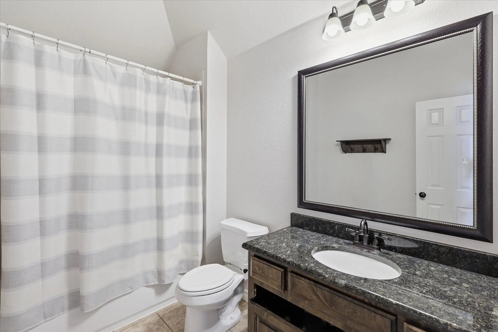 Bathroom featuring a stone-finish countertop with an undermount oval sink, dark-toned cabinetry, large-format tile flooring, and a framed wall mirror with integrated three-bulb vanity lighting