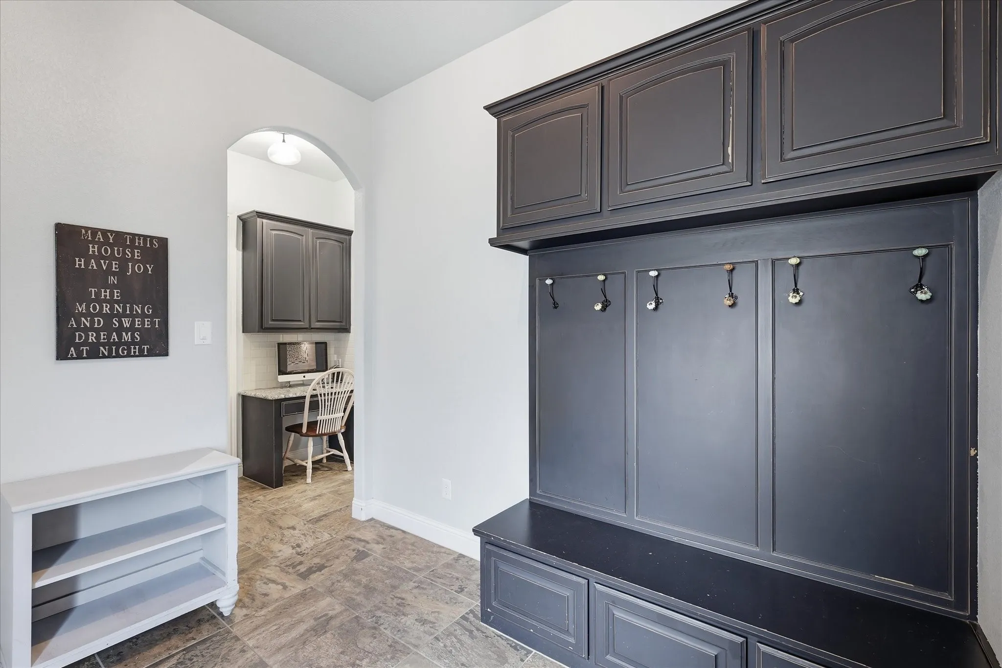 Built-in charcoal cabinetry with recessed panels, multi-hook coat rack, integrated bench seating, and an arched entryway leading to a dedicated workspace with stone-surface desk