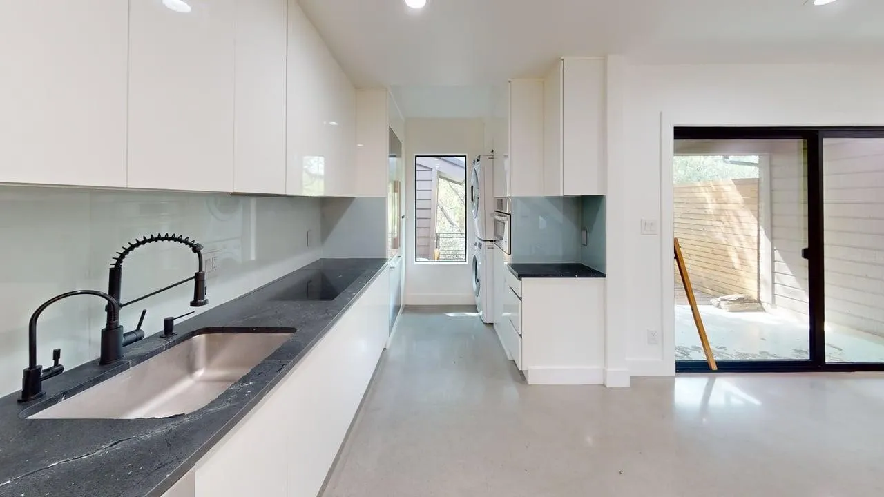 Modern kitchen featuring high-gloss white cabinetry, dark stone countertops, stainless steel undermount sink with a matte black arched faucet, and large-format sliding glass doors