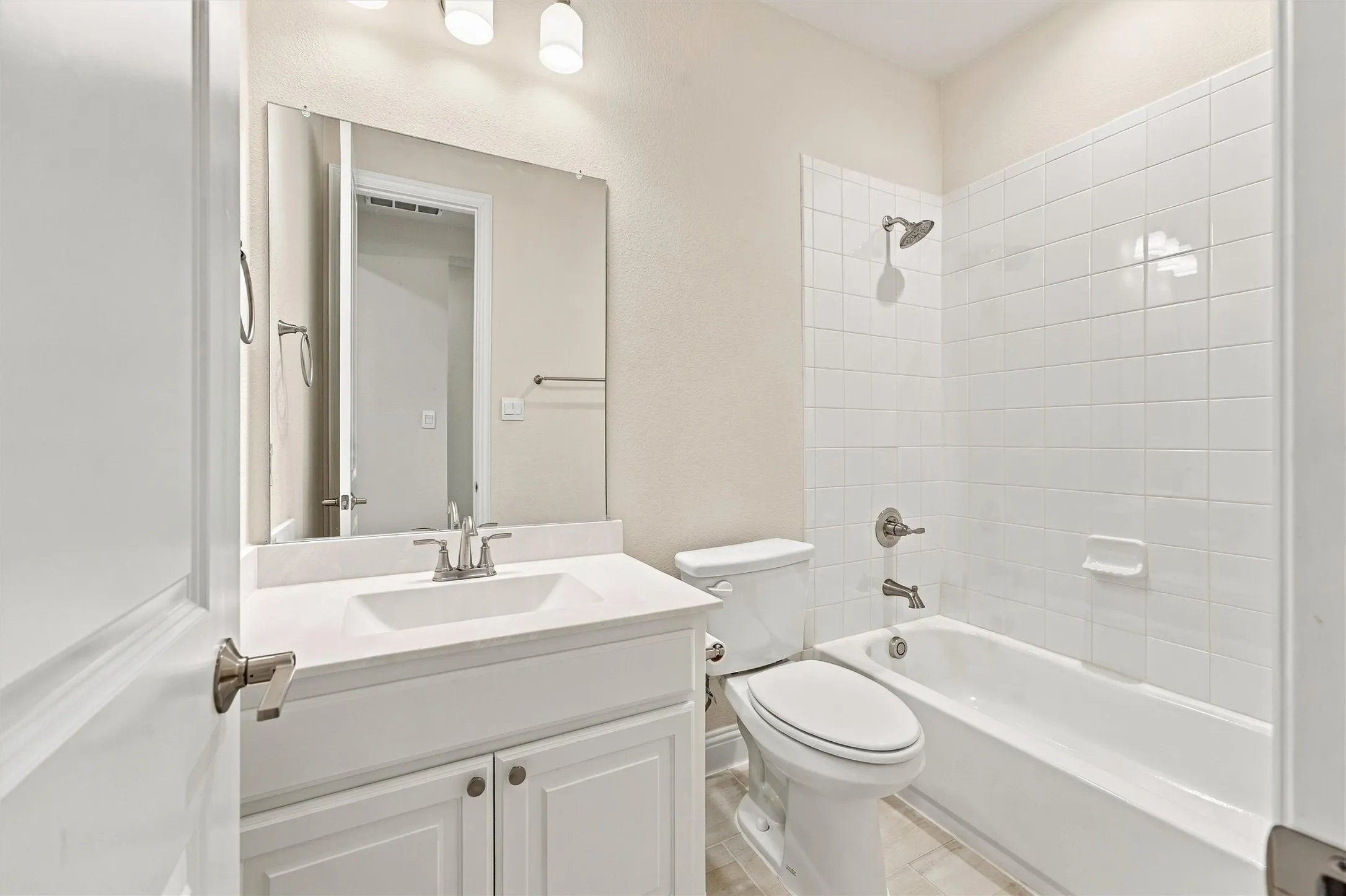 Bathroom featuring a vanity with white cabinetry, a rectangular sink, a wall-mounted mirror, a tub-shower combination with white tile surround, and brushed nickel hardware