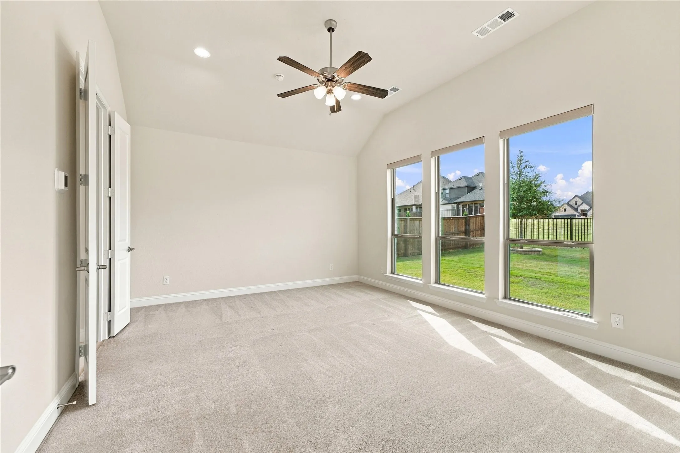 Vaulted ceiling, three large windows, neutral carpeting, and a multi-blade ceiling fan with integrated lighting