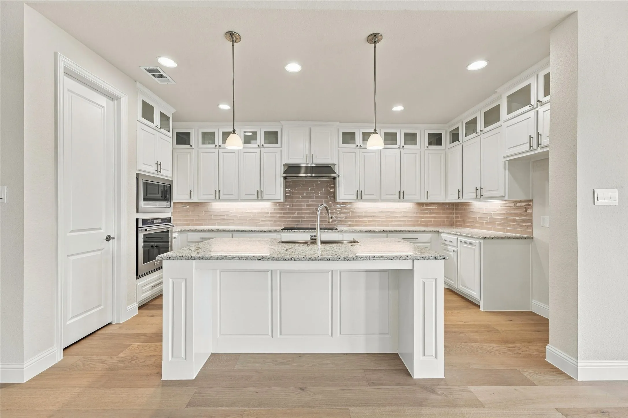 Kitchen featuring white cabinetry with glass-front uppers, a central island with stone-finish countertops, stainless steel built-in double ovens, linear tile backsplash, and wood-finish flooring