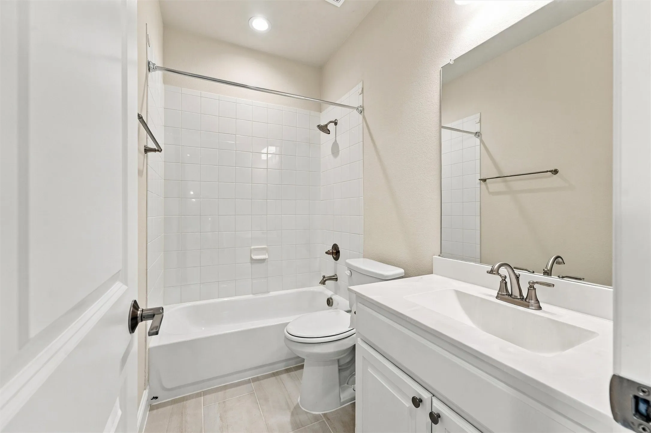 Bathroom featuring a white vanity with a composite countertop, an undermount sink, a tub-shower combination with white wall tile, and light-toned tile flooring
