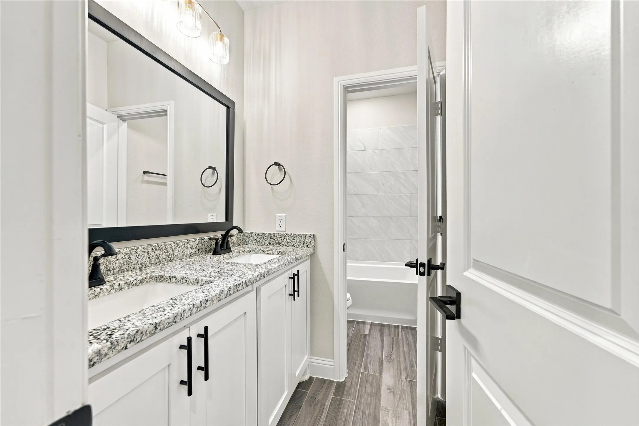 Dual-sink vanity with speckled stone countertop, matte black hardware, rectangular undermount sinks, framed vanity mirror, and wood-finish flooring