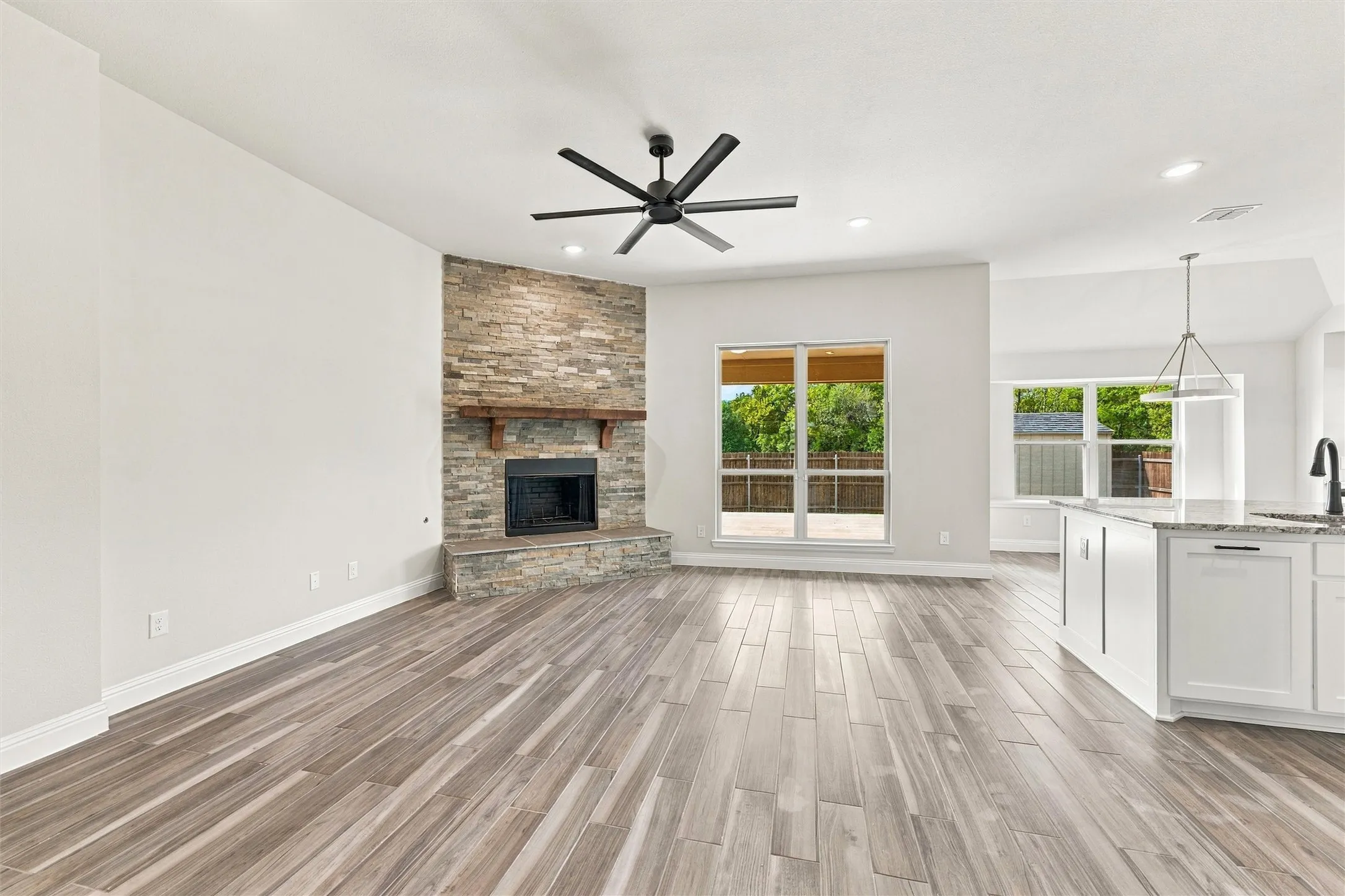 Open-concept living space featuring wood-finish flooring, a floor-to-ceiling stone-veneer fireplace with a heavy timber mantel, a multi-blade ceiling fan, and recessed lighting