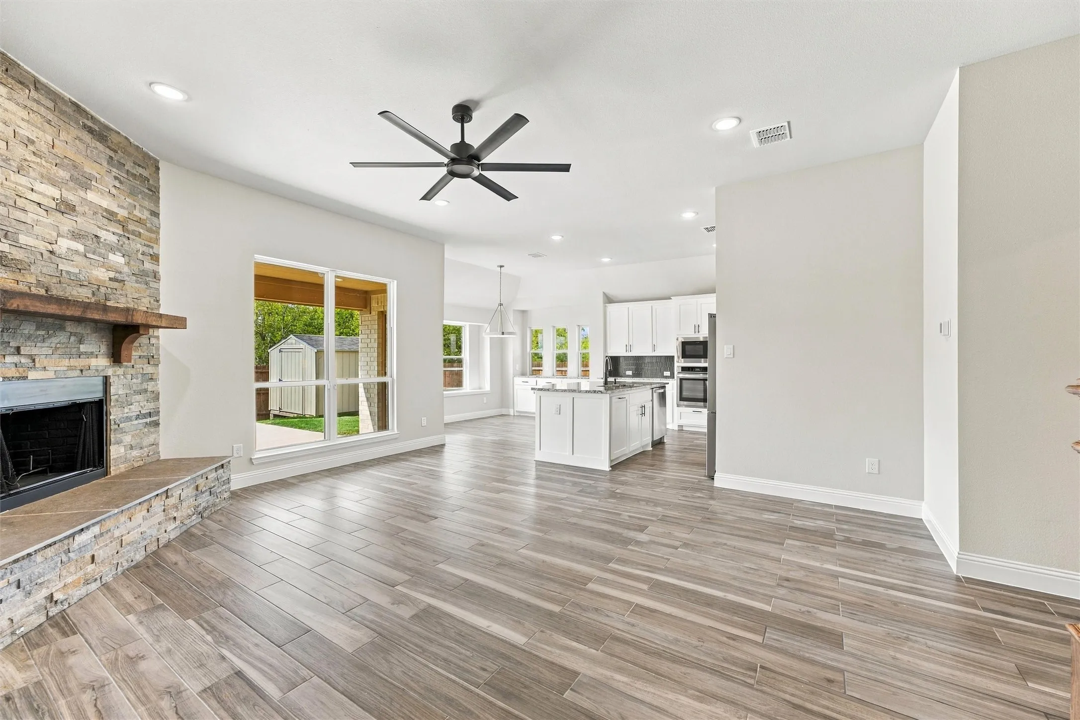 Stone-clad fireplace with a wood mantel, wood-finish flooring, ceiling-mounted fan, recessed lighting, and white kitchen cabinetry with a center island