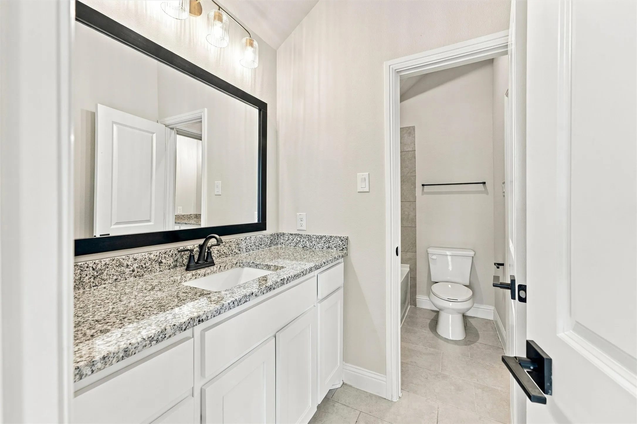 White cabinetry vanity with stone-finish countertop, undermount sink, matte black faucet, full-width framed mirror, and three-light vanity fixture