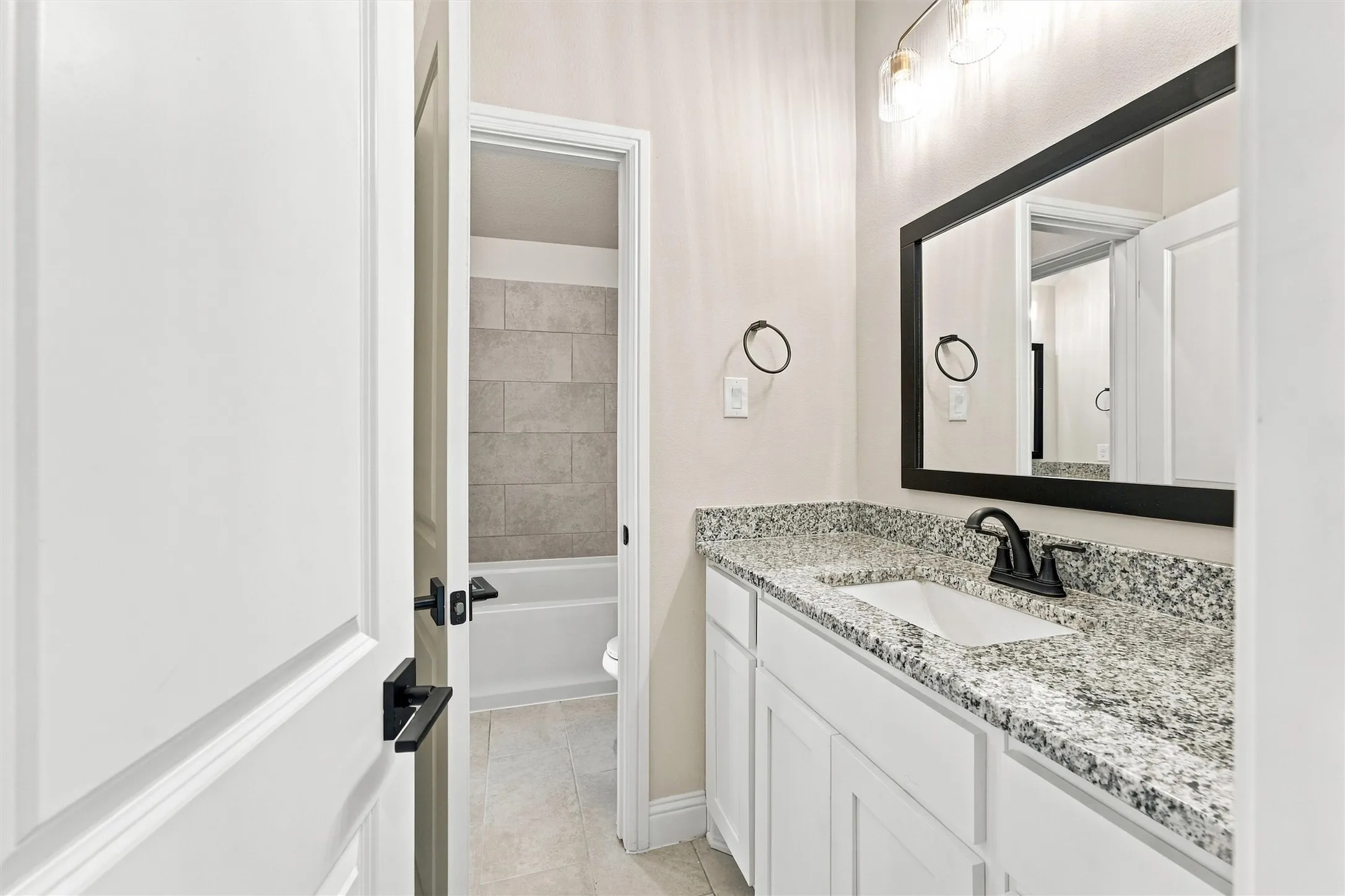Bathroom vanity featuring a stone-patterned countertop, under-mount sink with matte black faucet, shaker-style white cabinetry, framed black mirror, and three-bulb vanity lighting