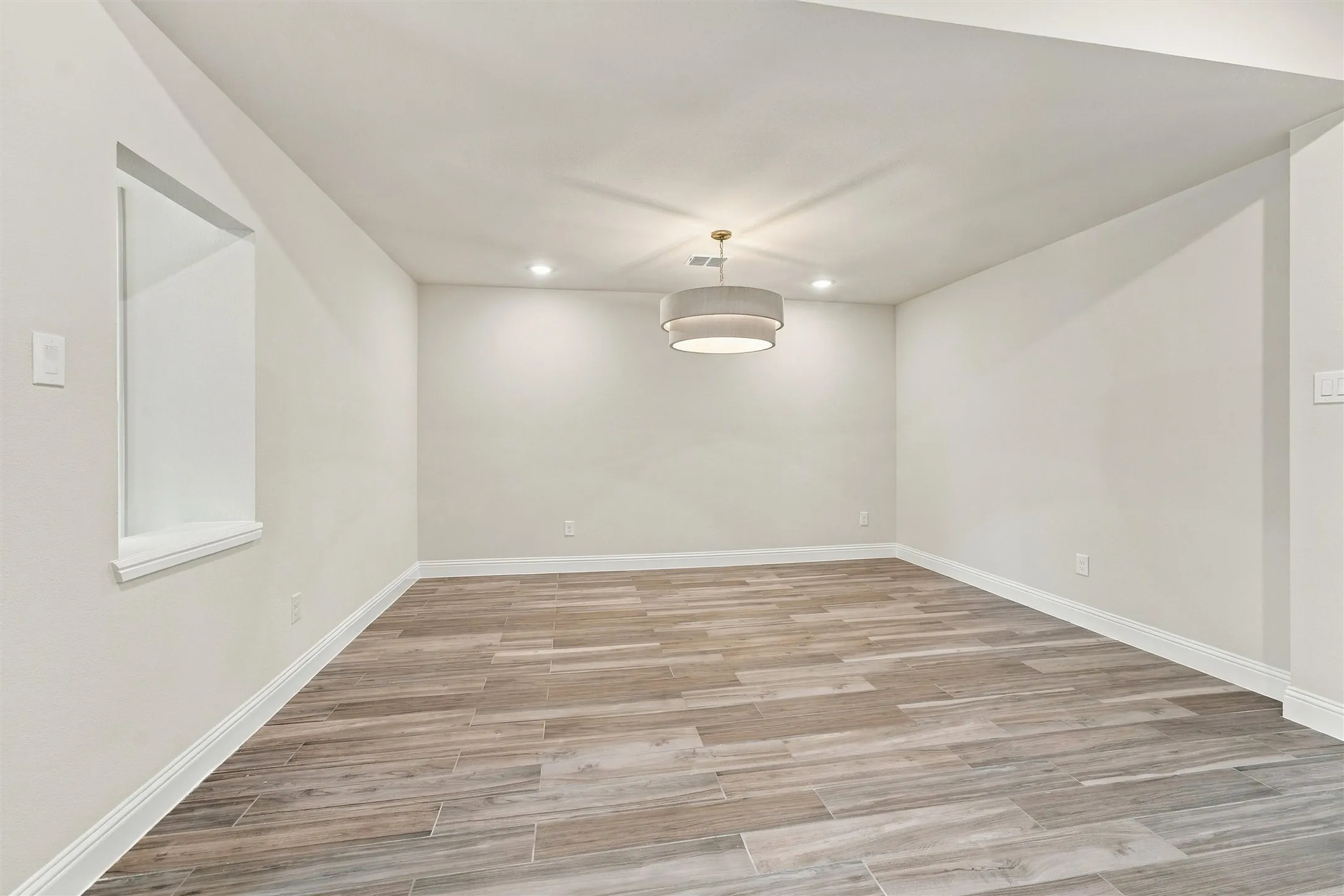 Neutral-toned interior space featuring wood-finish flooring, a tiered drum pendant light, recessed ceiling lighting, and a built-in wall niche