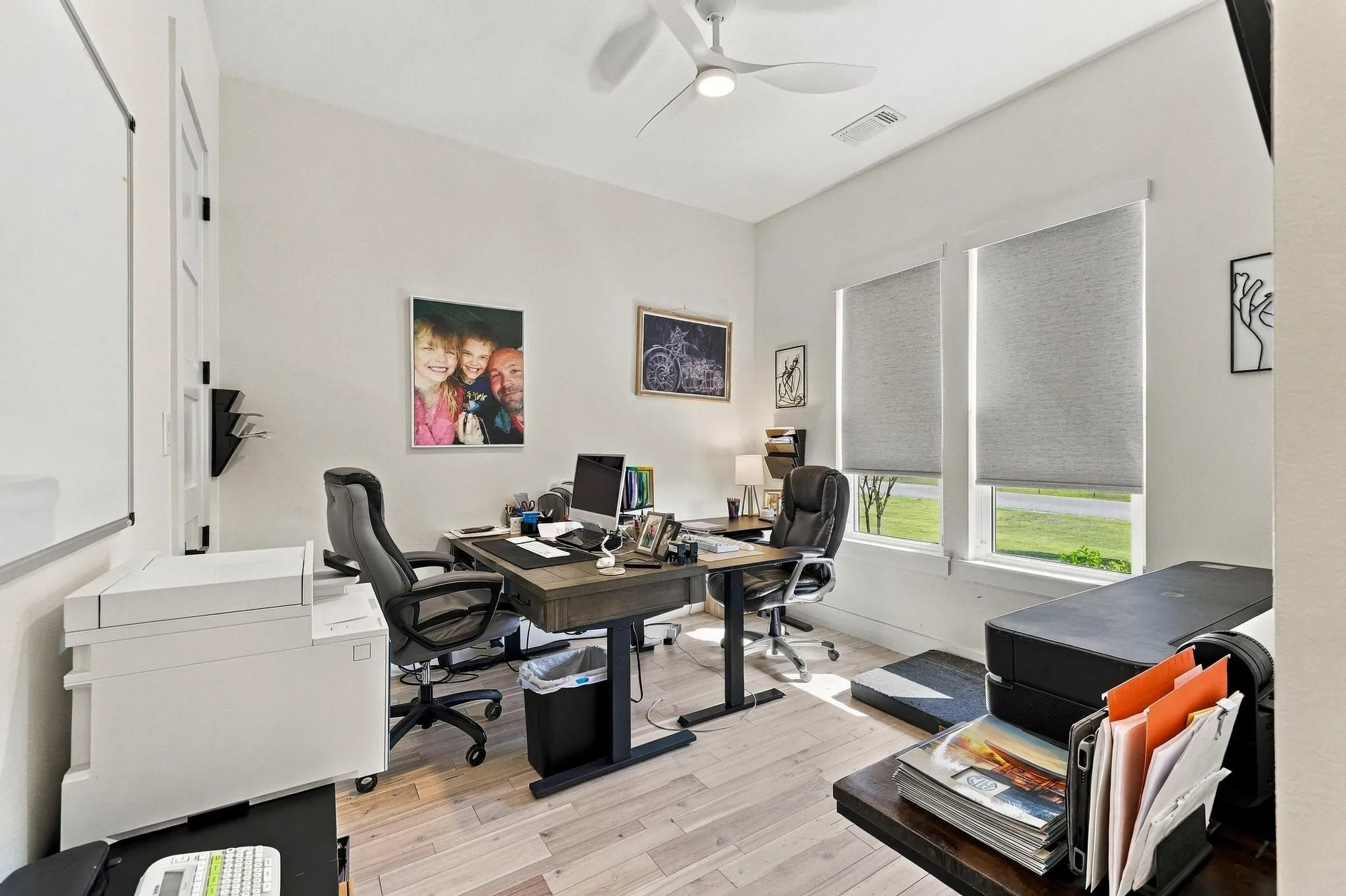 Neutral-toned room featuring wood-finish flooring, a white three-blade ceiling fan with integrated lighting, and dual windows with fitted shades