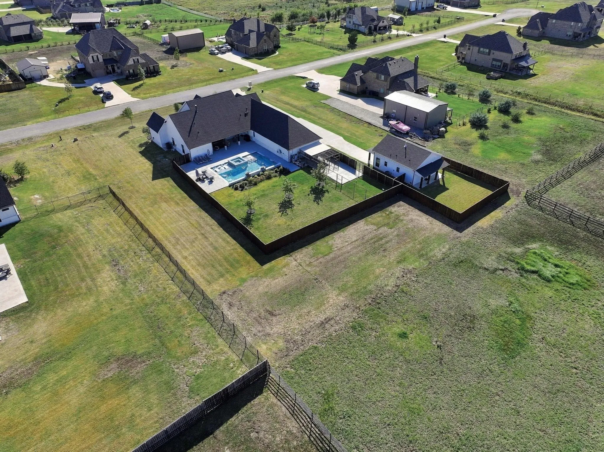 Aerial perspective of a multi-structure property featuring a swimming pool, covered patio, fenced grounds, and a detached building