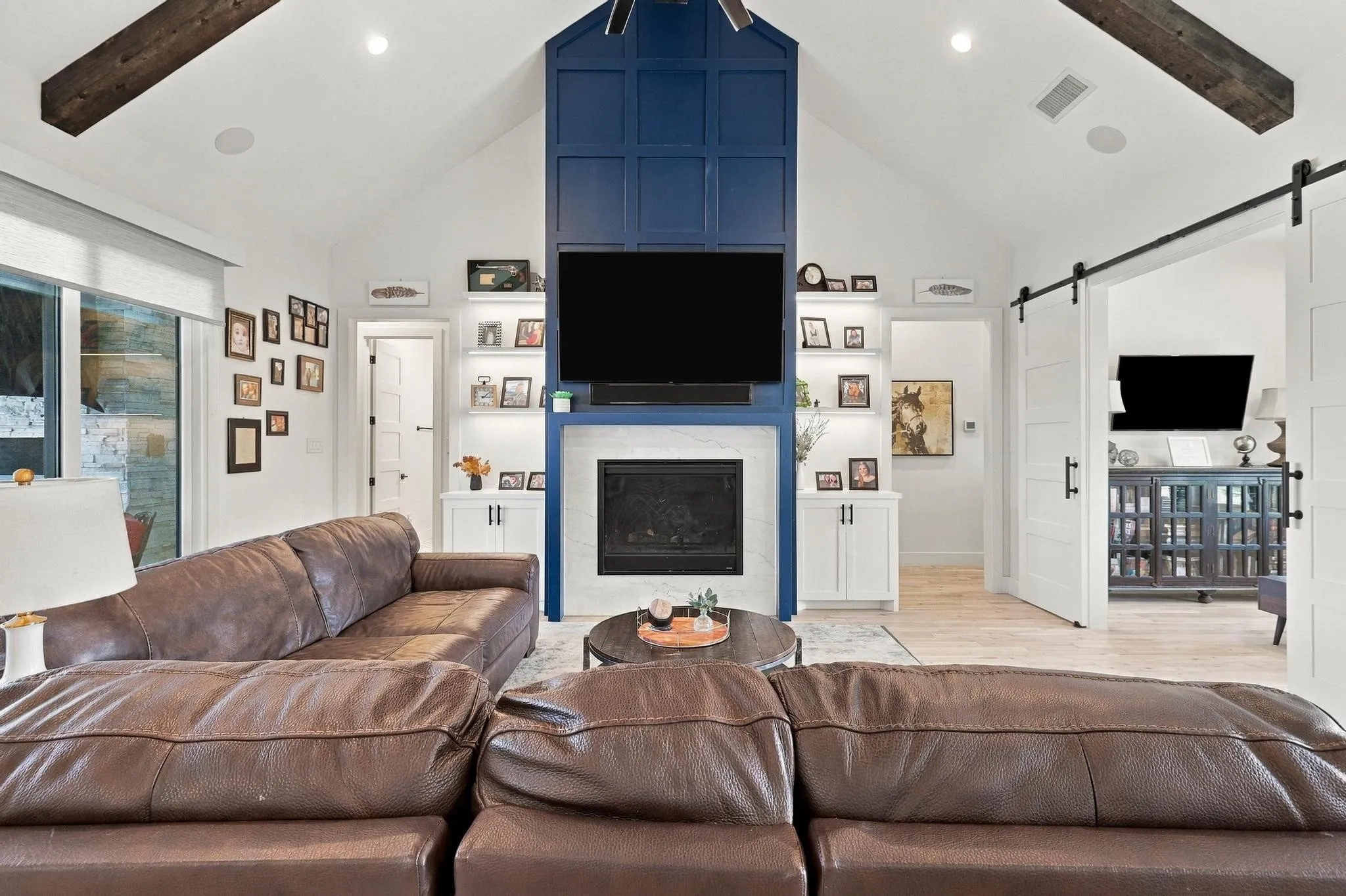 Vaulted ceilings with exposed wood beams, fireplace with a custom floor-to-ceiling blue paneled surround, built-in display shelving, and white sliding barn doors with matte black hardware