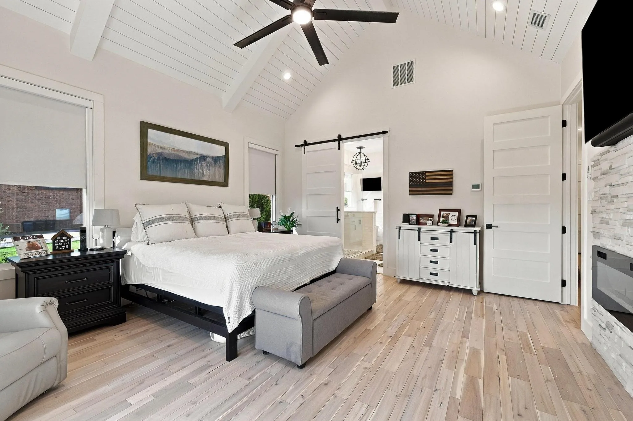 Vaulted ceiling with shiplap detailing, light-toned wood-finish flooring, stone-surround built-in fireplace, sliding barn door, and ceiling fan
