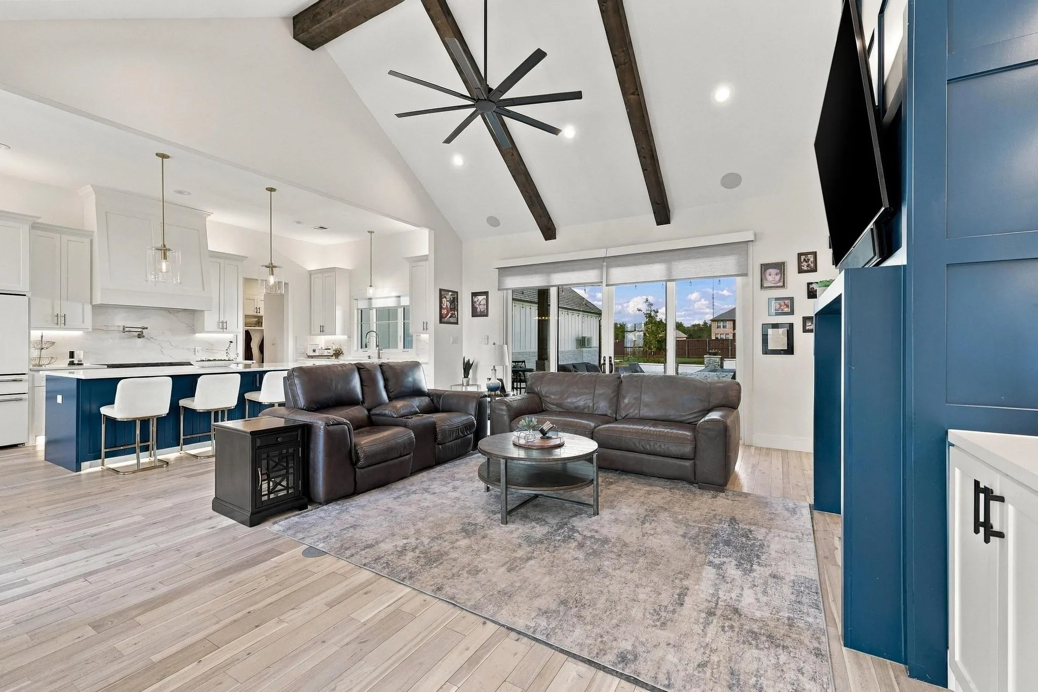 Vaulted ceiling with exposed wood beams, wood-finish flooring, floor-to-ceiling accent wall, kitchen featuring a center island with bar seating, and large windows with recessed lighting