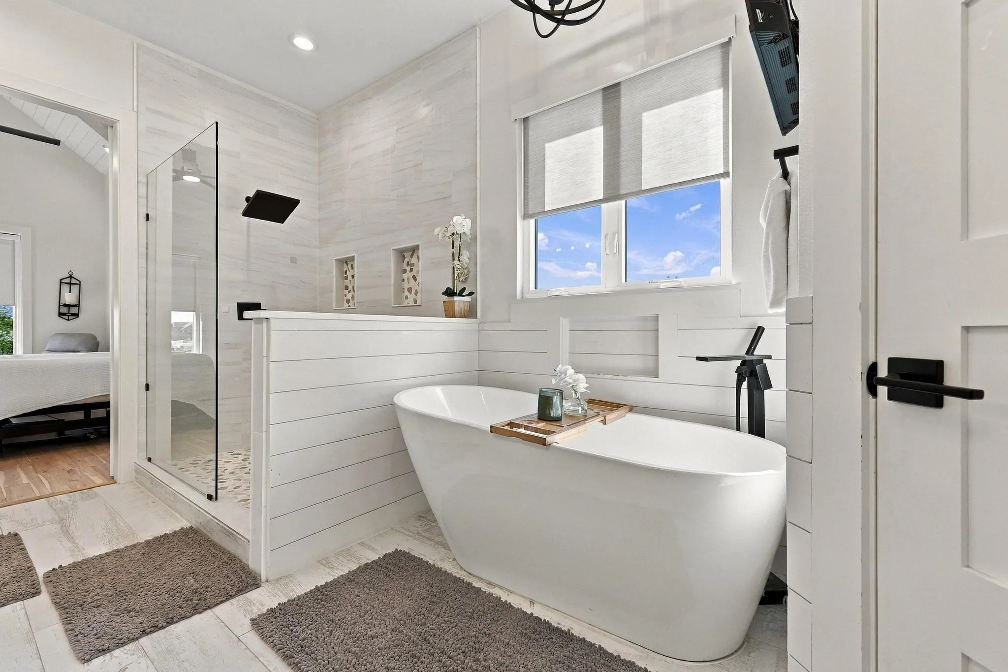 Freestanding soaking tub with a matte black floor-mounted faucet, walk-in shower featuring a frameless glass enclosure and rainfall showerhead, horizontal shiplap wall accents, and recessed wall shelving