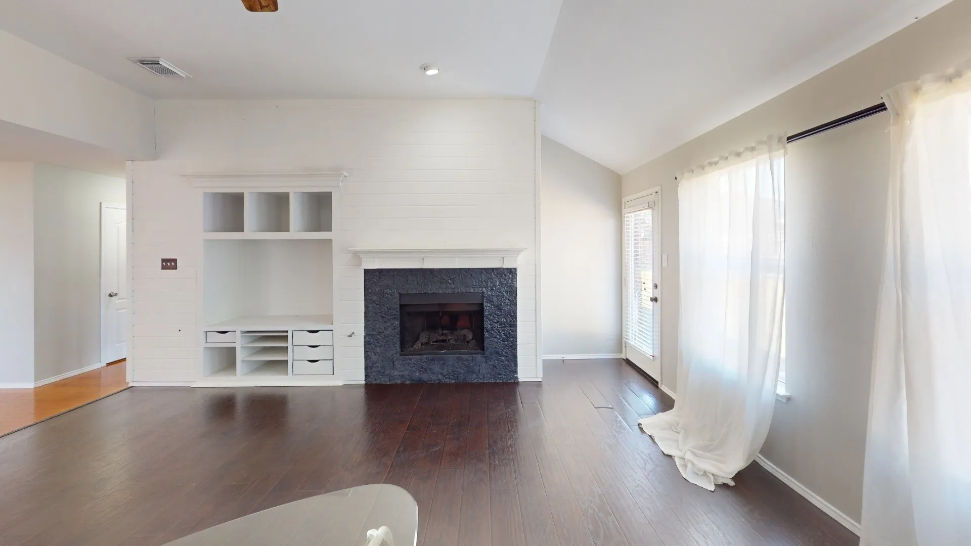 Living area featuring wood-finish flooring, a vaulted ceiling, a floor-to-ceiling painted brick fireplace with a textured stone surround, and built-in shelving with integrated storage drawers