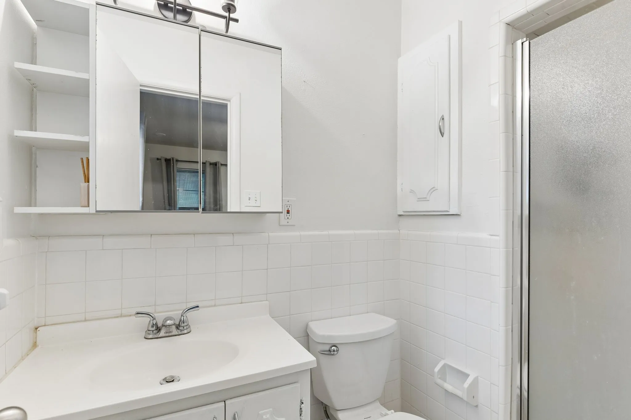 Bathroom featuring white wall tiling, a vanity with an integrated sink, a mirrored medicine cabinet with open shelving, and a glass-enclosed shower stall